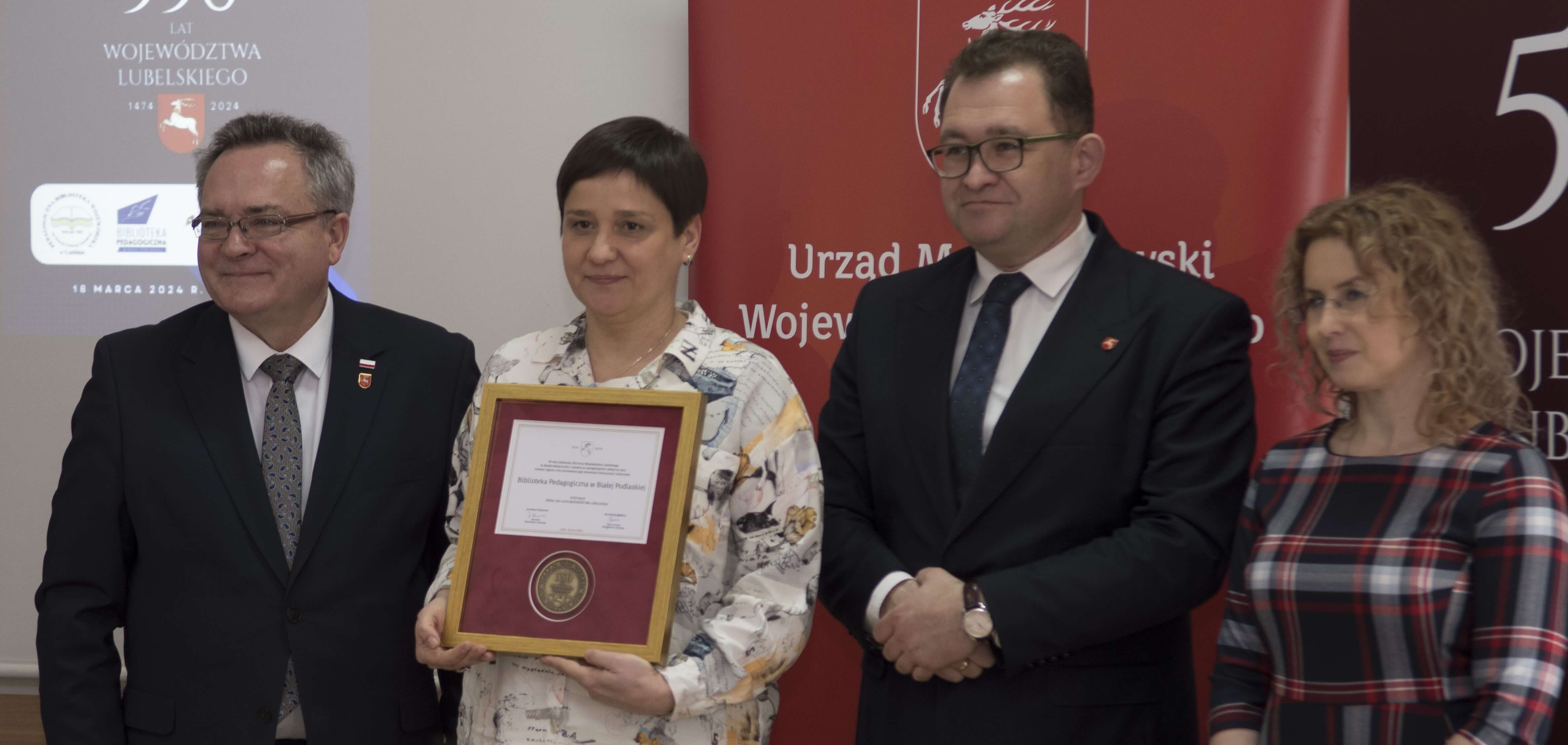 Wicedyrektor Biblioteki Pedagogicznej w Białej Podlaskiej prezentuje medal otrzymany z okazji jubileuszu 550-lecia województwa lubelskiego. Po obu stronach wicedyrektor stoją dwaj przedstawiciele Urzędu Marszałkowskiego w Lublinie.